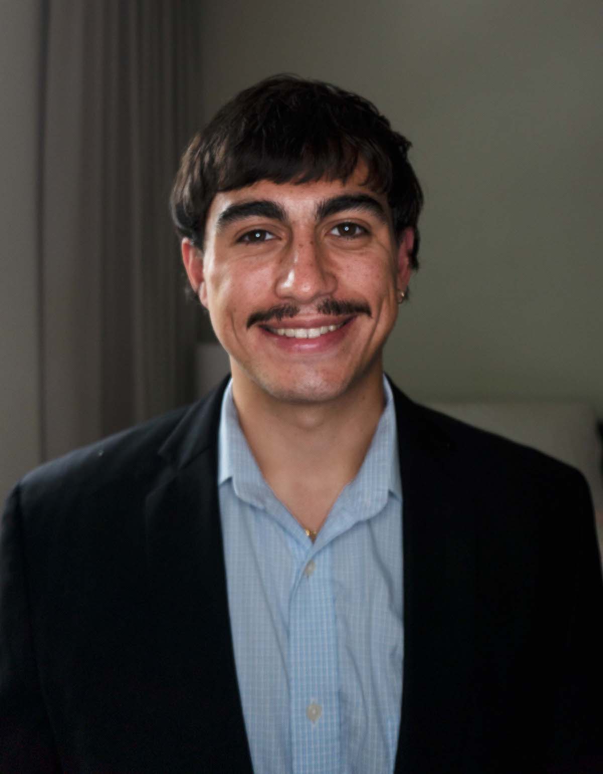 Headshot of a 20 year old man. He is smiling brightly and is wearing business professional clothing. His hair is choppy and he wears a mustache and an earring